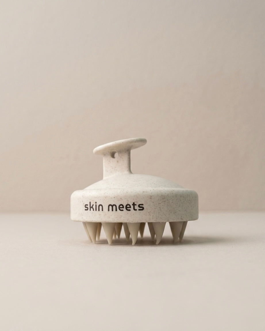 A round, textured off-white hair massager brush with a handle, featuring the text 'skin meets' printed in black on the front, set against a minimalist beige background.