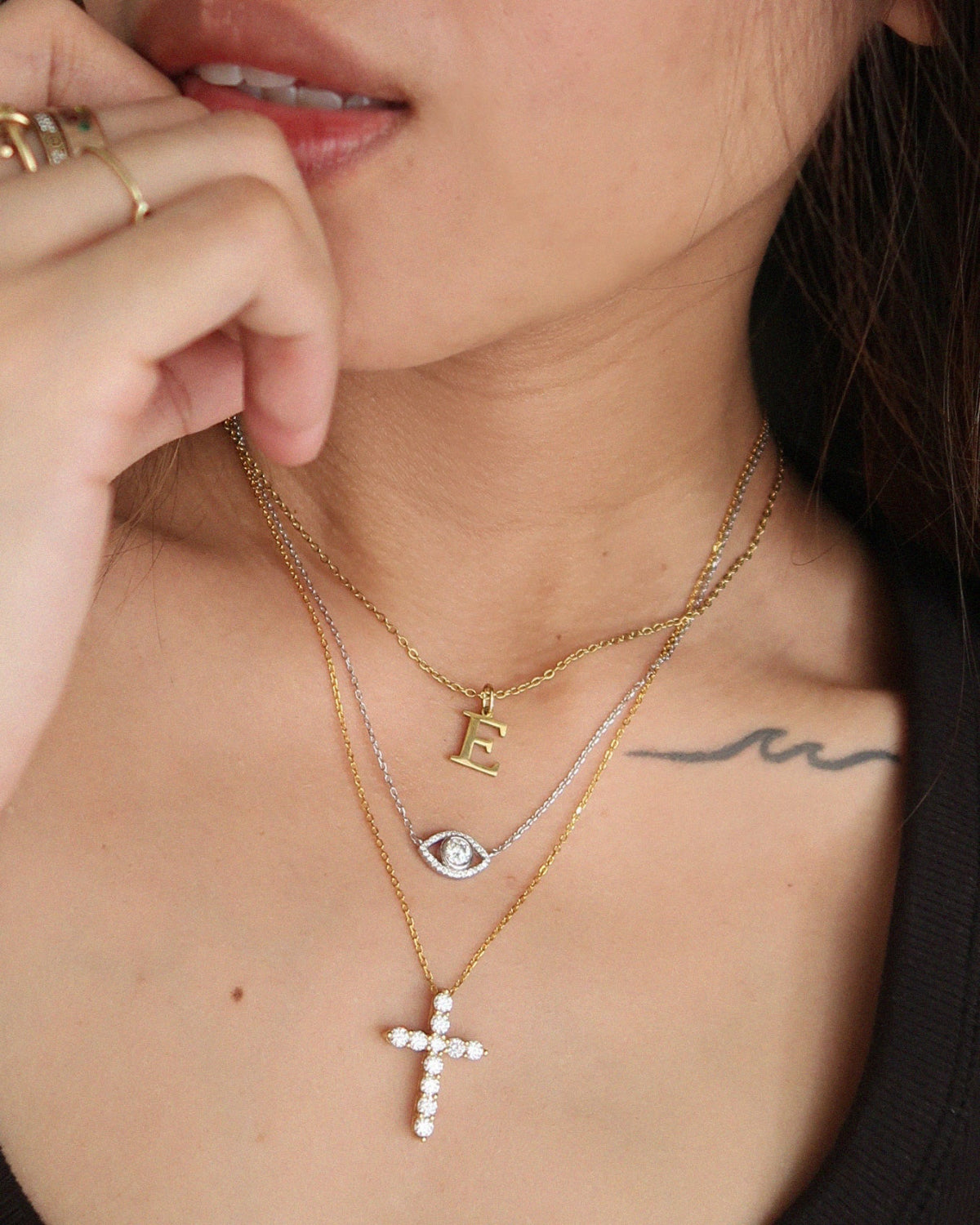 Close-up of a person wearing multiple necklaces with a cross pendant, letter 'E' pendant, and eye pendant.